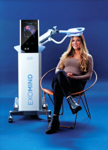 Paradise Medical Maui, Woman seated next to Exomind TMS therapy device. Brain scan displayed on screen. Exomind branding visible.