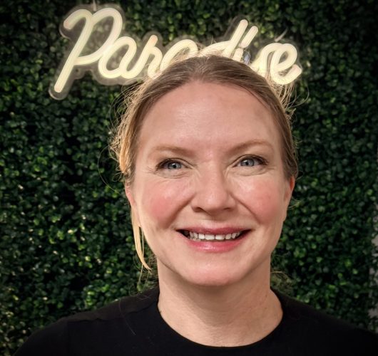 Paradise Medical Maui, Smiling woman in front of a green wall with a neon "Paradise" sign.